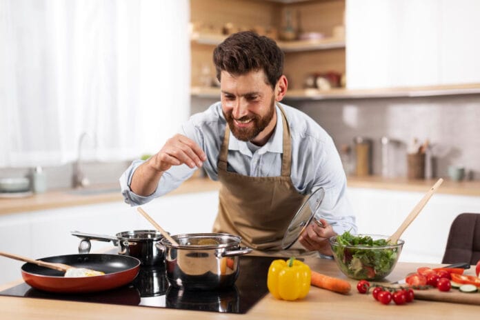 cheerful-adult-european-bearded-man-chef-apron-prepares-dinner-family-salts-soup-modern-kitchen